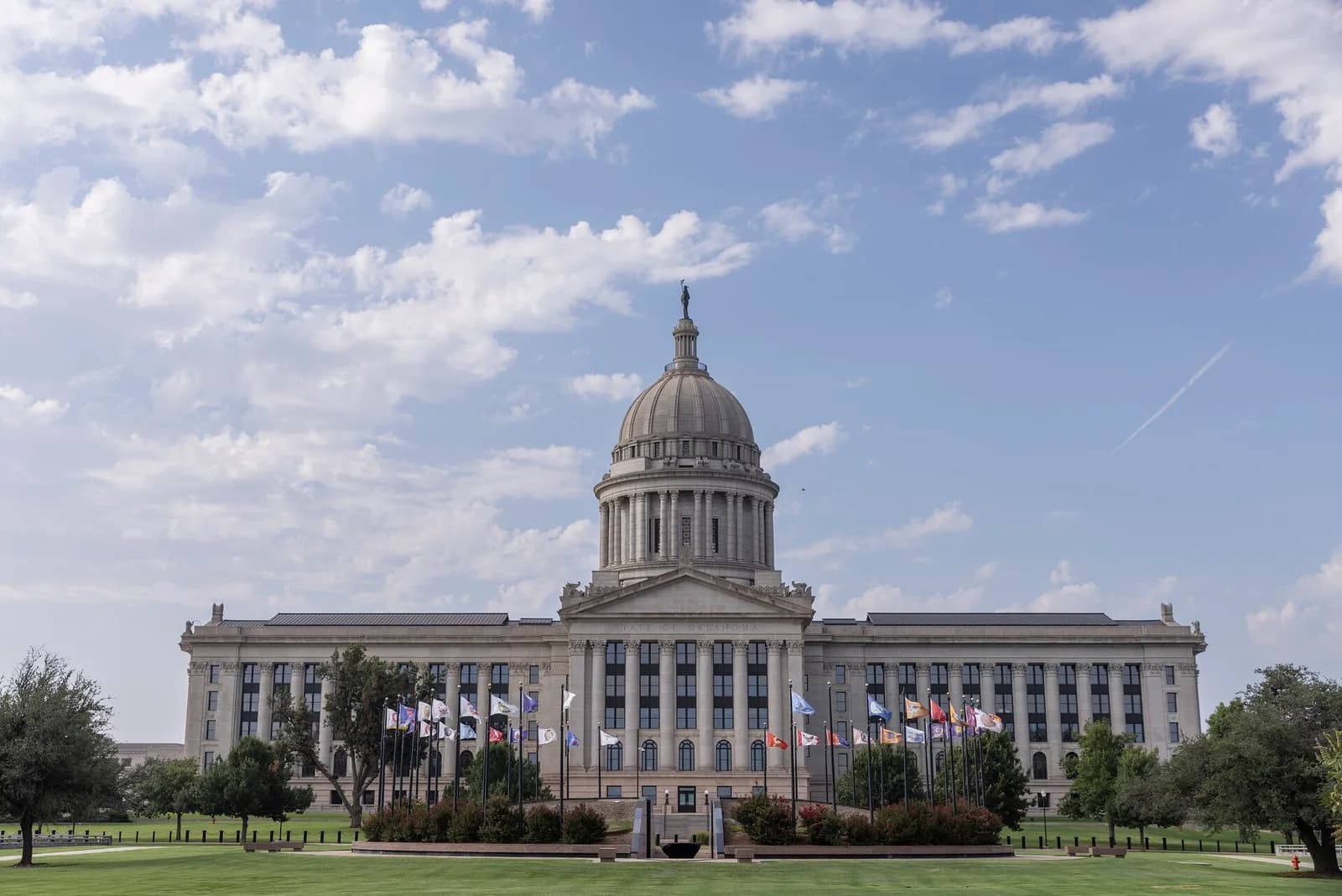 A large, classical-style building with a central dome and a statue on top stands under a partly cloudy sky. Numerous flags are displayed on poles in front of the building, which is surrounded by green lawns and some trees.