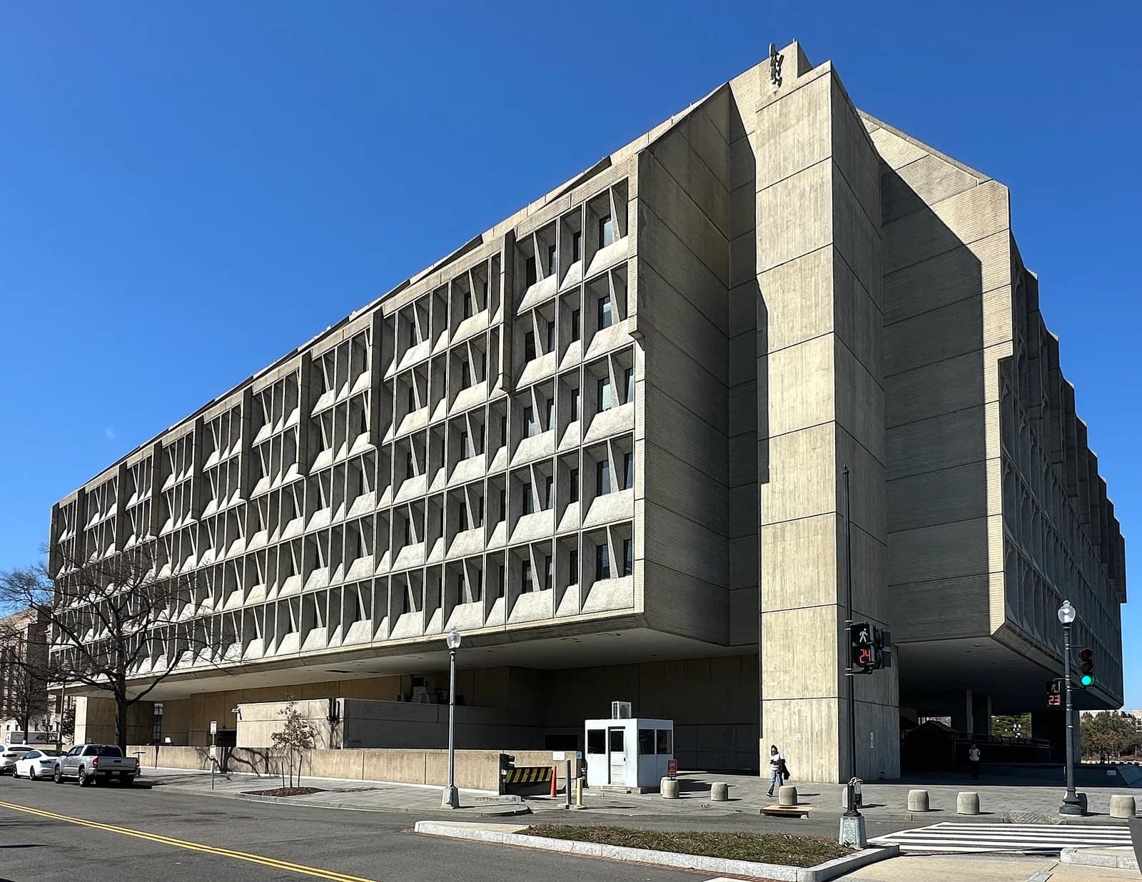 A large, brutalist concrete building with a distinctive facade of recessed, angular windows stands under a clear blue sky. The building is set back from a street with a crosswalk, a small guard booth, and a few people walking nearby.