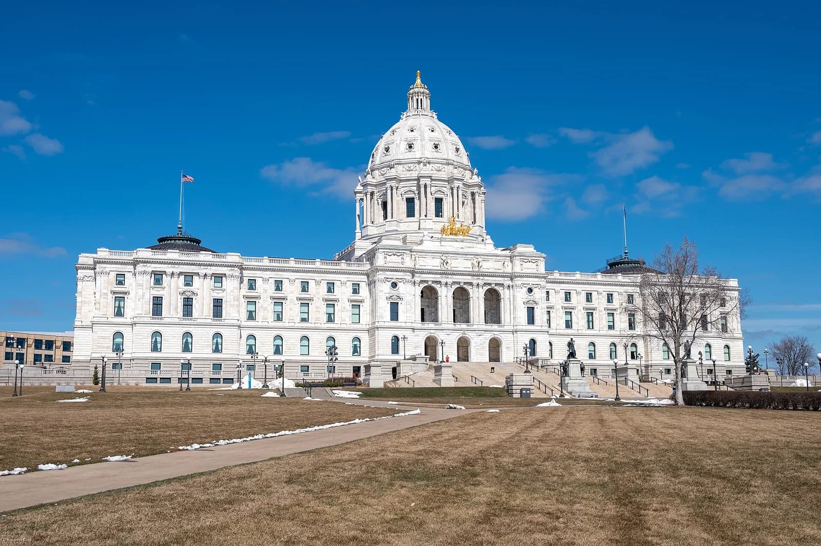 A large, white neoclassical building with a prominent dome and a flag flying on a clear day. The building is surrounded by a brown grassy lawn with patches of snow and a paved path leading towards it.