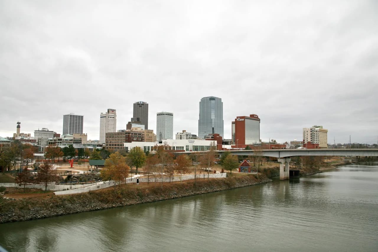 A cityscape view shows a river in the foreground with a bridge crossing it. On the far bank, a park with autumn trees leads to a skyline of various tall buildings under a cloudy sky.