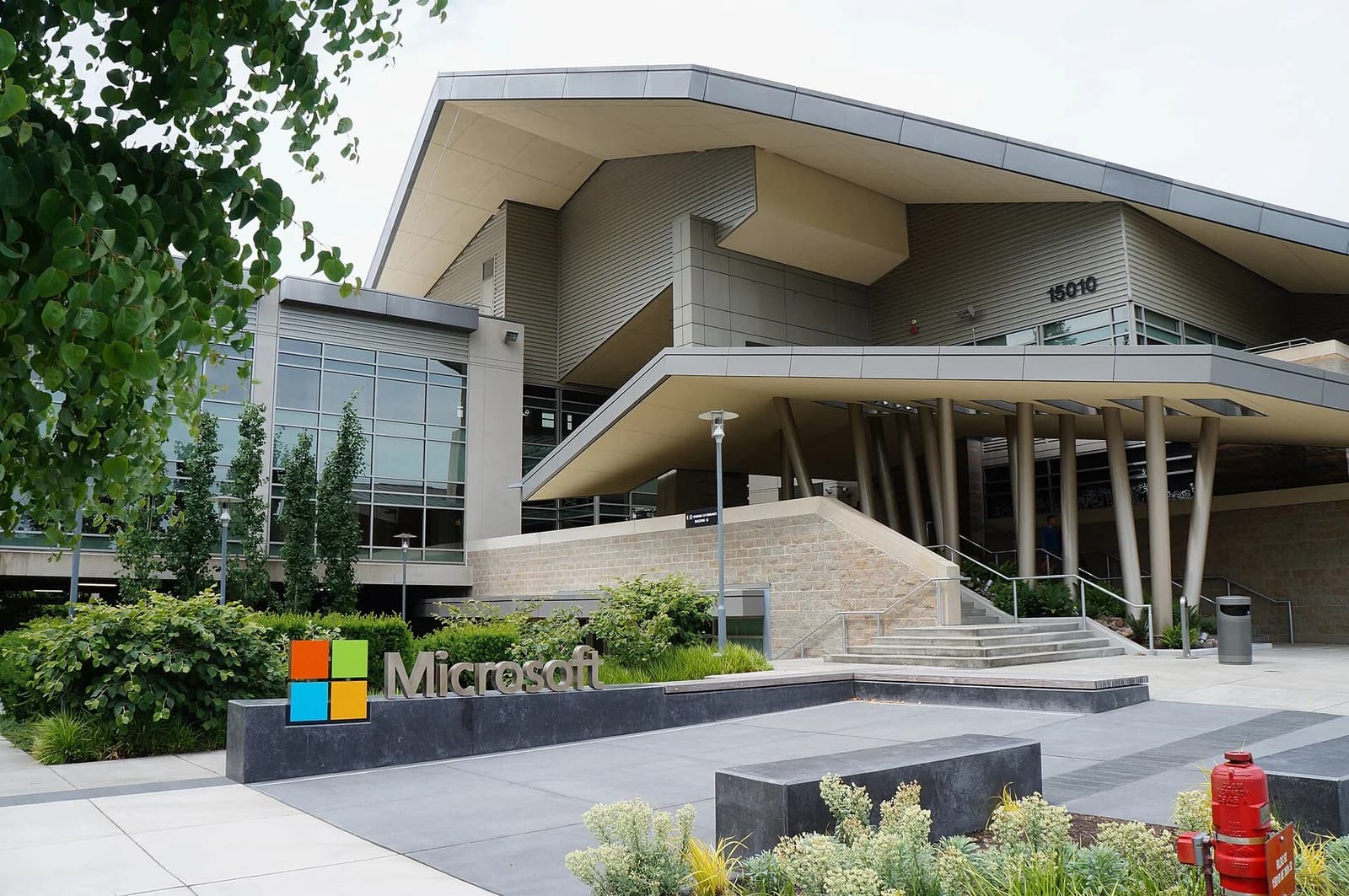 A modern building with a large, angular roof and glass windows is visible. A sign with a colorful logo and the word "Microsoft" is in the foreground, surrounded by landscaping.
