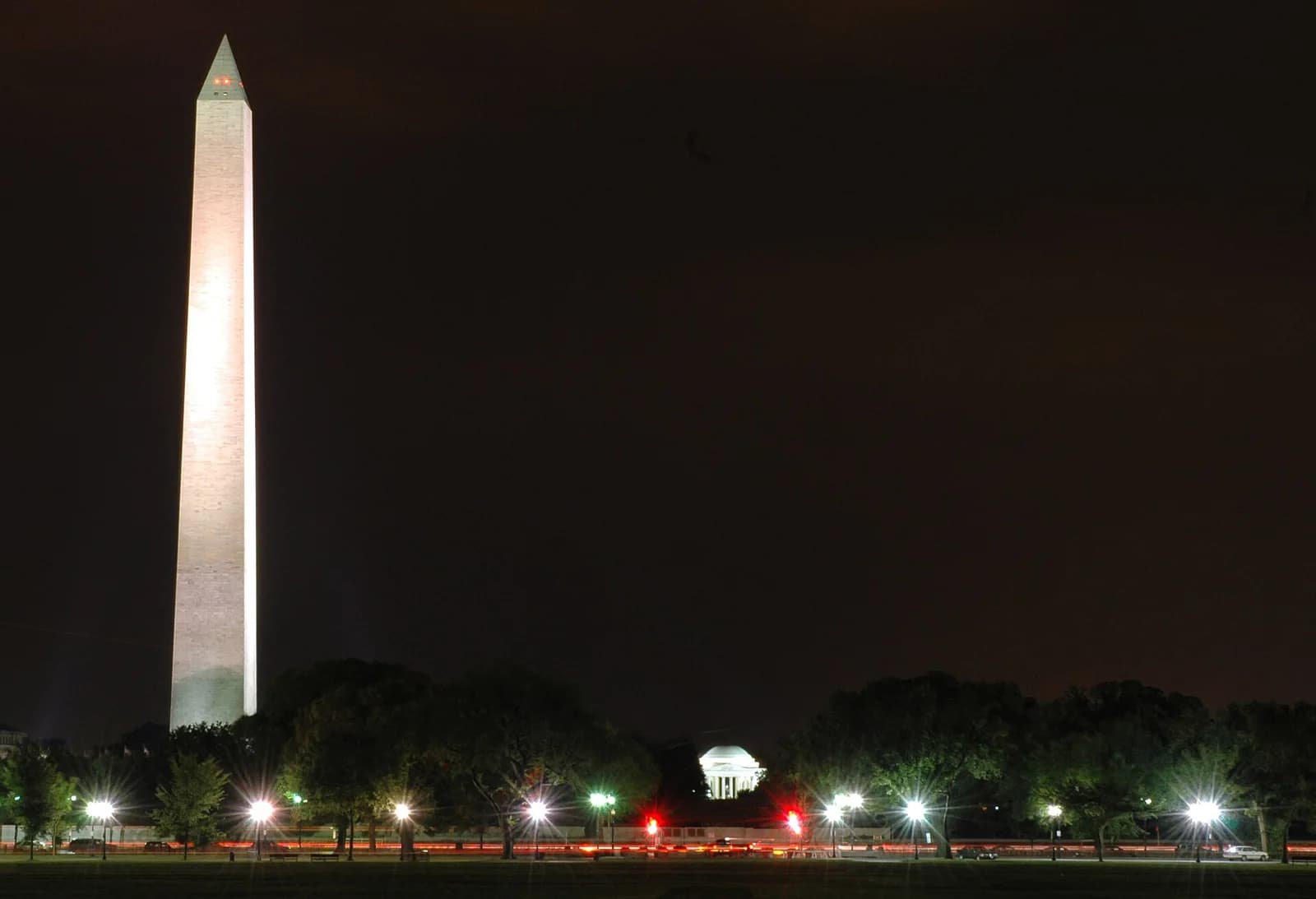 A tall, illuminated white obelisk stands prominently on the left against a dark night sky. In the distance, a smaller, domed white building is visible behind a line of trees and streetlights, with red and white light trails from traffic on a road in the foreground.