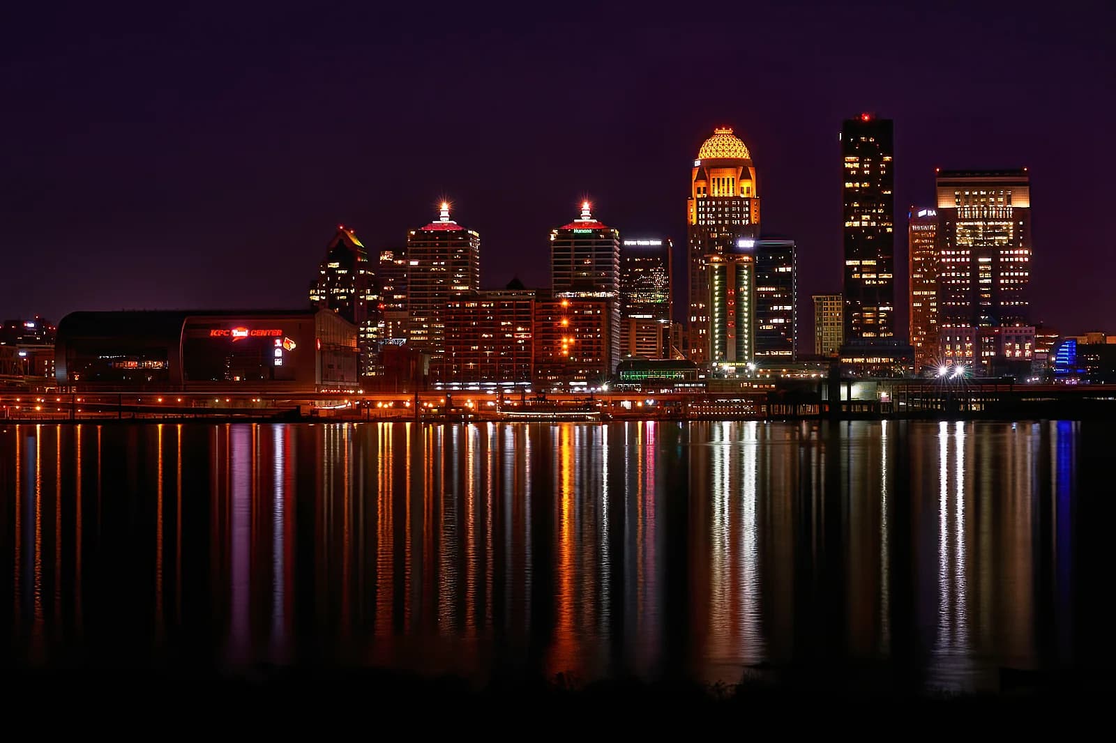 A nighttime cityscape shows a collection of illuminated buildings across a body of water, with their lights reflecting on the water's surface. The sky is dark, and the buildings feature various architectural styles and heights.