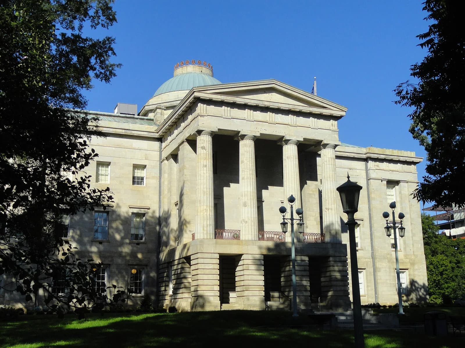 A large, light-colored stone building with a prominent portico supported by columns and a green dome on top is visible. The building is surrounded by green grass and trees, with lampposts in the foreground.