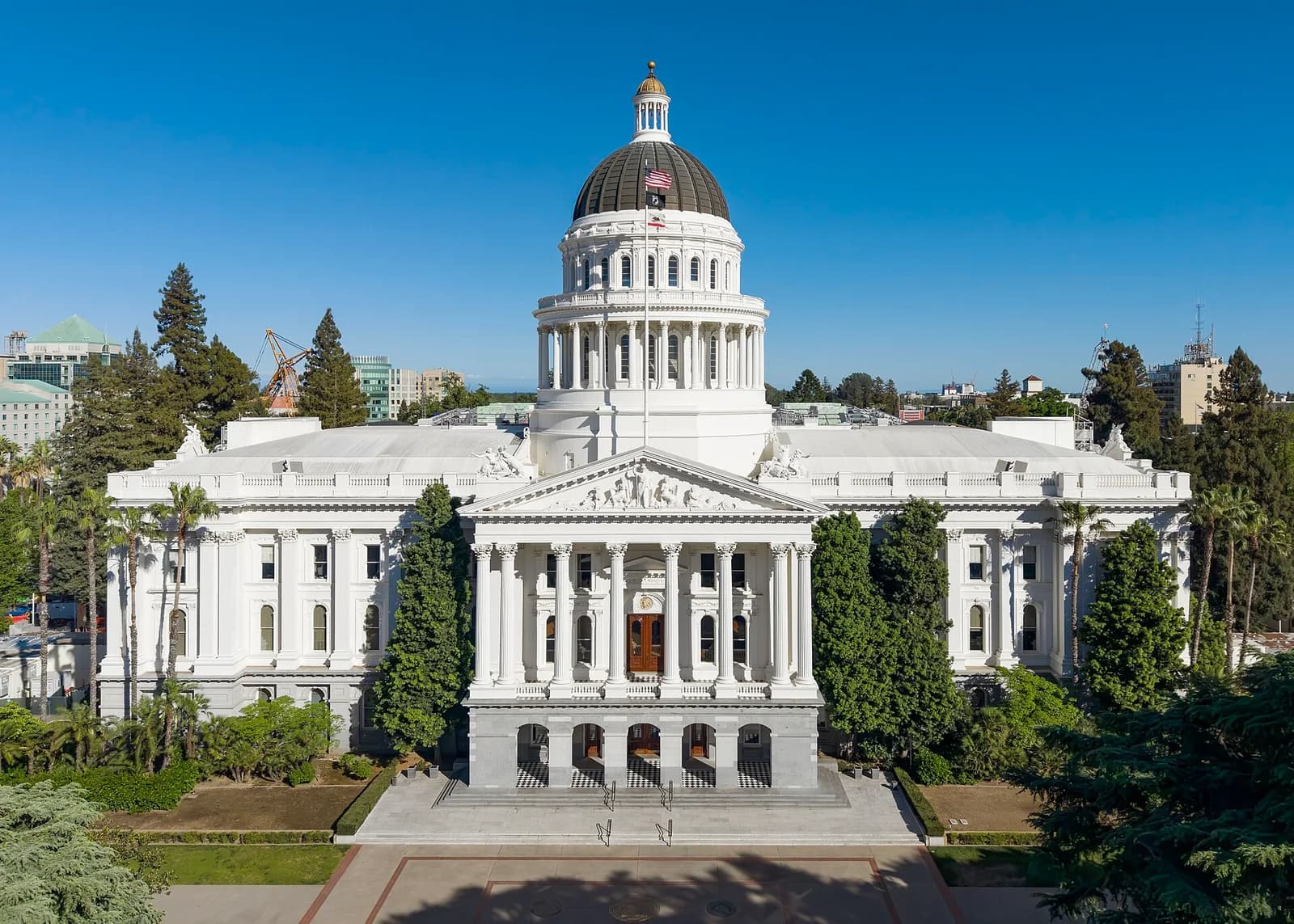 An aerial view shows a large white neoclassical building with a prominent dome and portico, surrounded by green trees and landscaping under a clear blue sky. The building features columns, ornate carvings, and a flag flying from the dome.