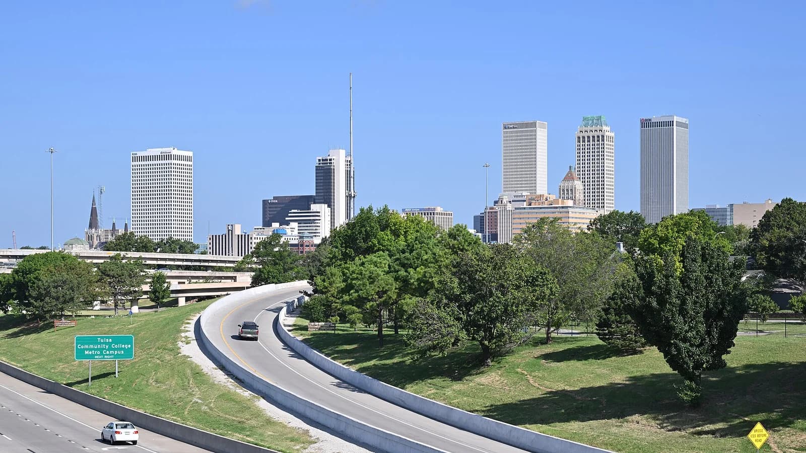 A cityscape view shows multiple tall buildings under a clear blue sky, with a multi-lane highway curving through the foreground. Green trees and grassy areas are visible between the highway and the distant buildings.