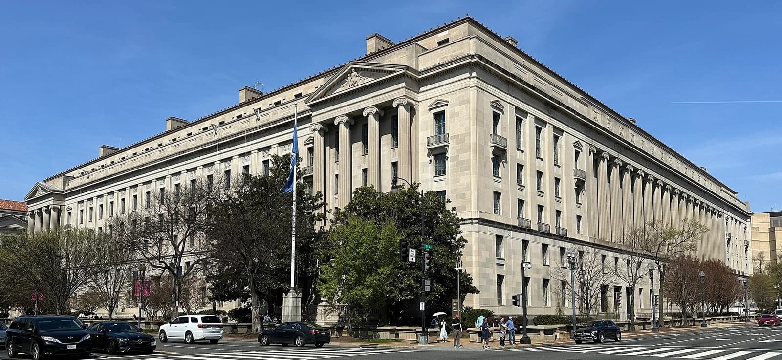 A large, light-colored stone building with classical architectural elements, including columns and a pediment, stands under a clear blue sky. Cars are parked and driving on the street in front of the building, and a few pedestrians are visible on the sidewalk.