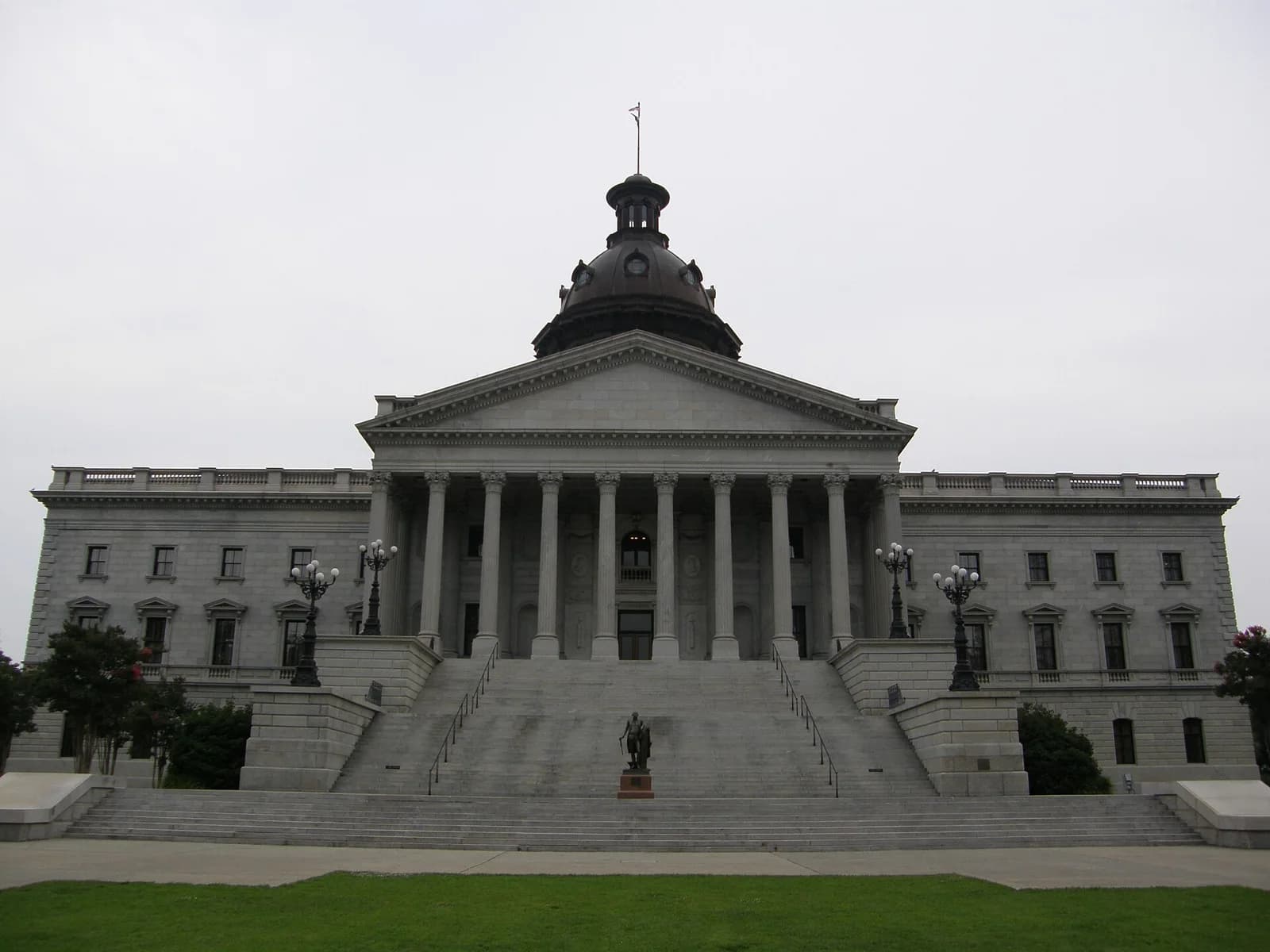A large, grey stone building with a central dome and a grand staircase leading up to a portico with columns. The building is surrounded by green grass and a few trees under an overcast sky.