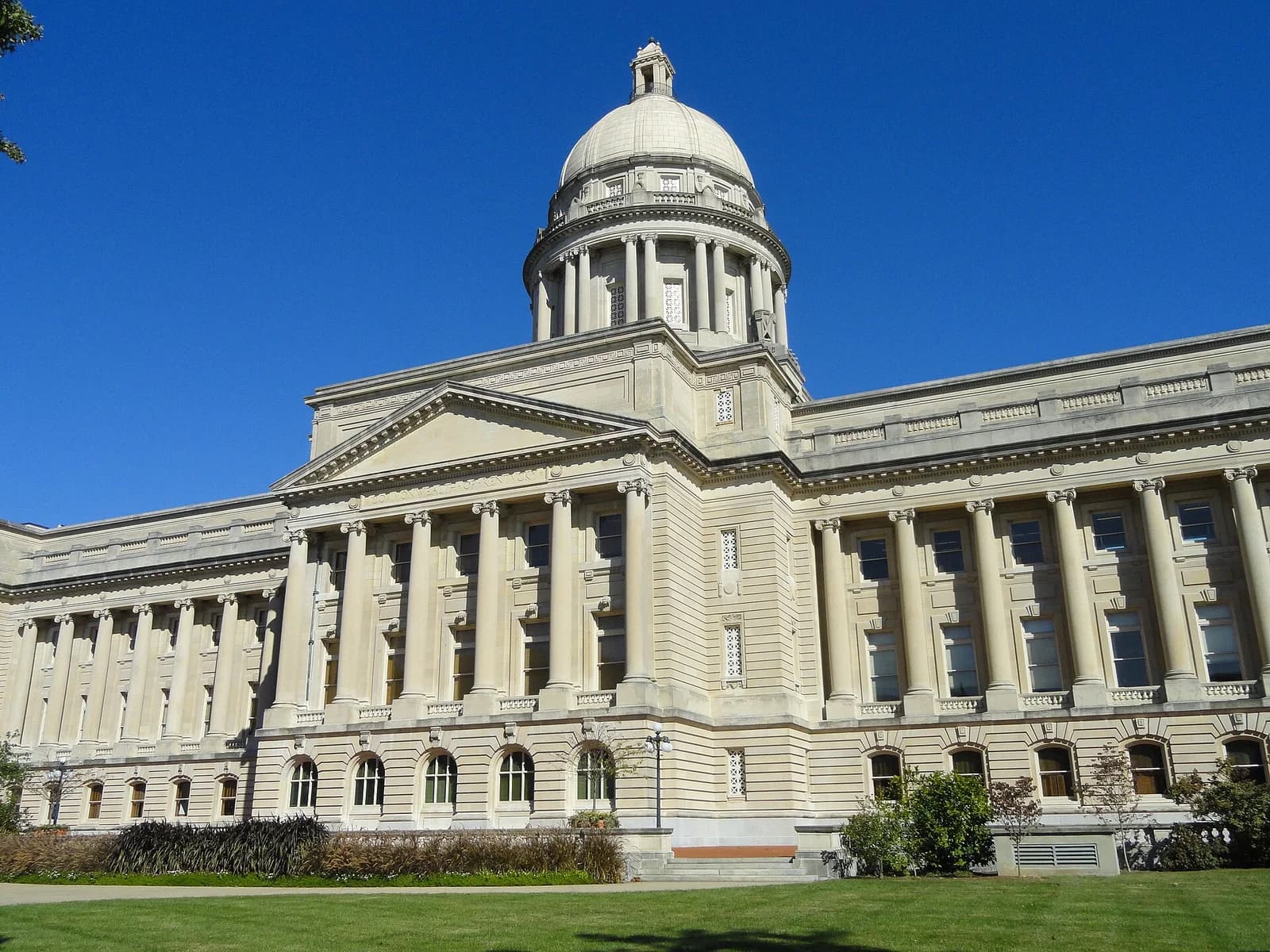 A large, ornate neoclassical building with a prominent dome and columns stands under a clear blue sky. The building is surrounded by green grass and some shrubbery in the foreground.