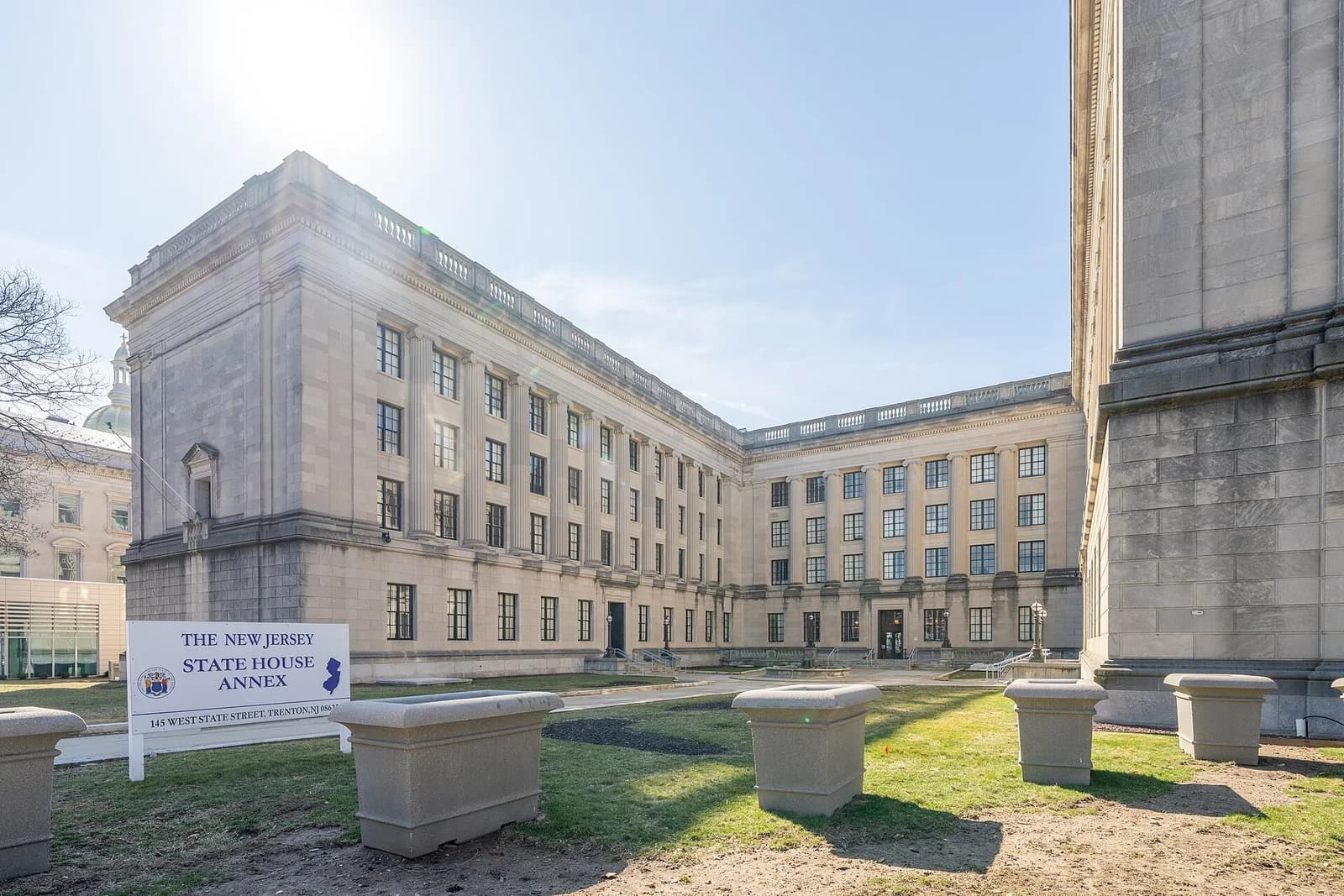 A large, light-colored stone building with many windows and columns forms an L-shape around a grassy area. A white sign with blue text stands in the foreground.