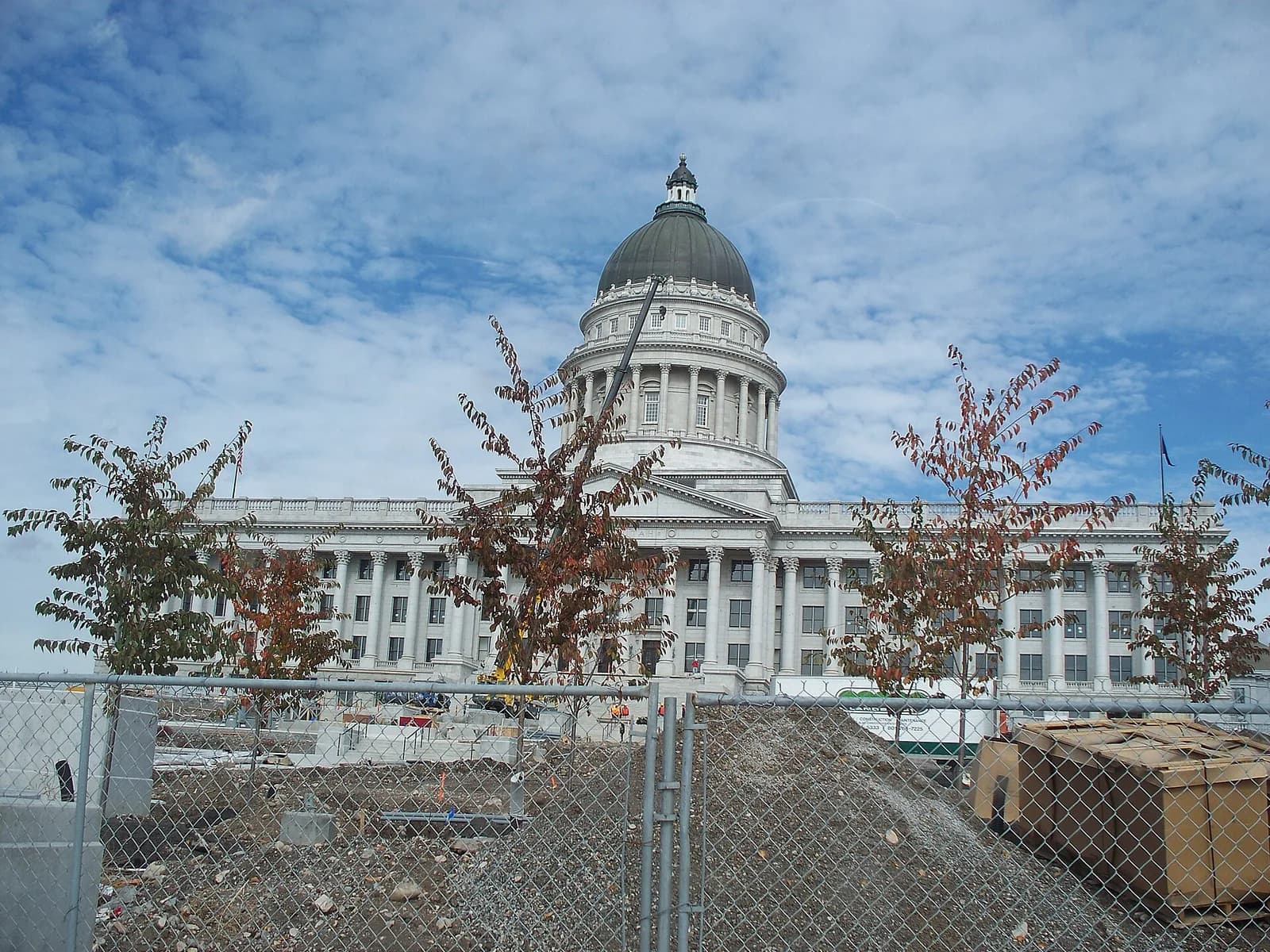 A large white neoclassical building with a prominent dome and columns is visible behind a chain-link fence and a construction site. The sky is partly cloudy with some trees in the foreground.