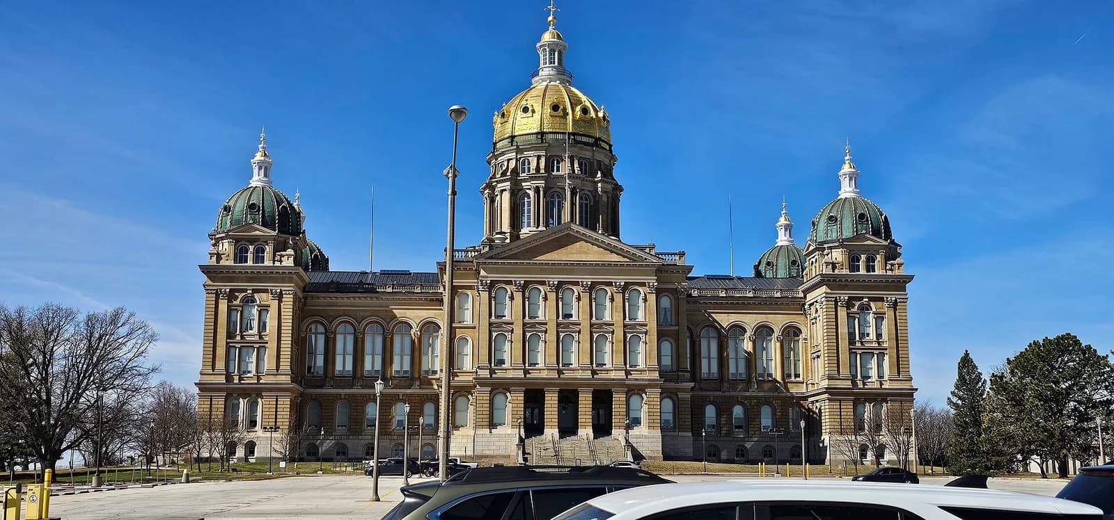 A large, ornate building with a prominent golden dome in the center and two smaller green domes on either side stands under a clear blue sky. The building has multiple windows and architectural details, with trees and parked cars visible in the foreground and around the perimeter.