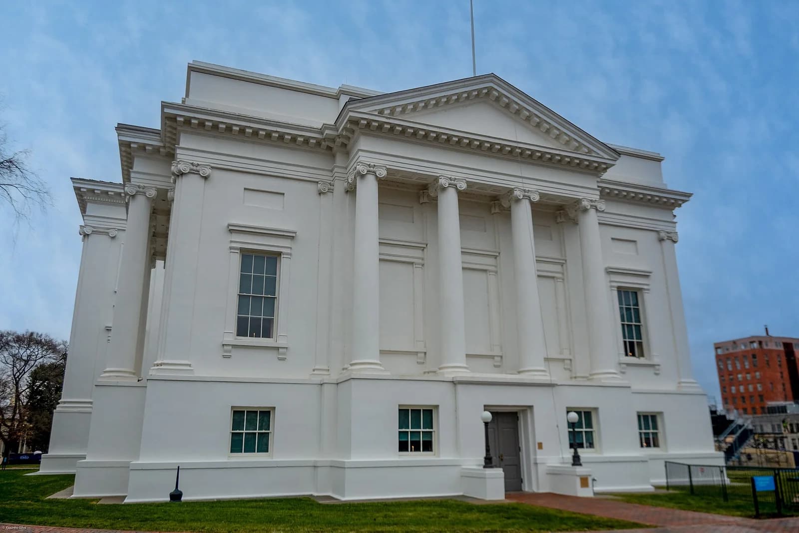 A large white neoclassical building with a prominent portico featuring Ionic columns and a pediment is shown under a cloudy sky. The building is surrounded by green grass and a brick pathway, with another building visible in the background to the right.