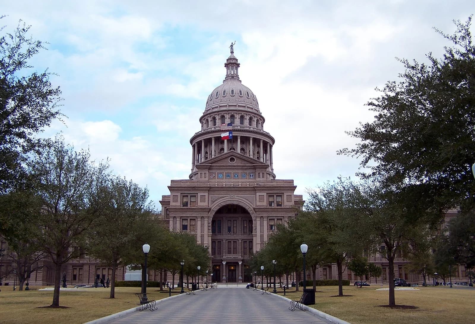 A grand, light-colored stone building with a large dome and a statue on top is visible at the end of a paved walkway. The building is surrounded by green lawns and numerous trees, with a cloudy sky overhead.