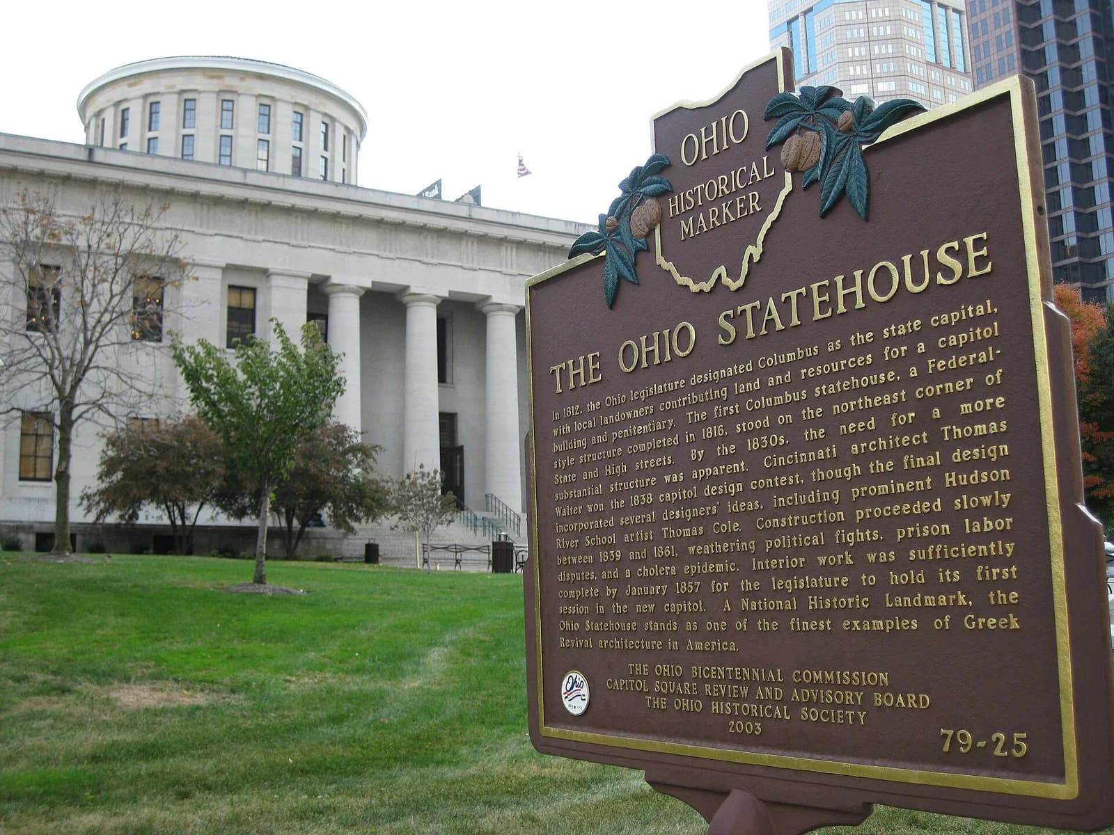 A large, light-colored stone building with a domed roof and columns is visible behind a brown historical marker. The building is surrounded by green grass and trees, with taller modern buildings in the background to the right.