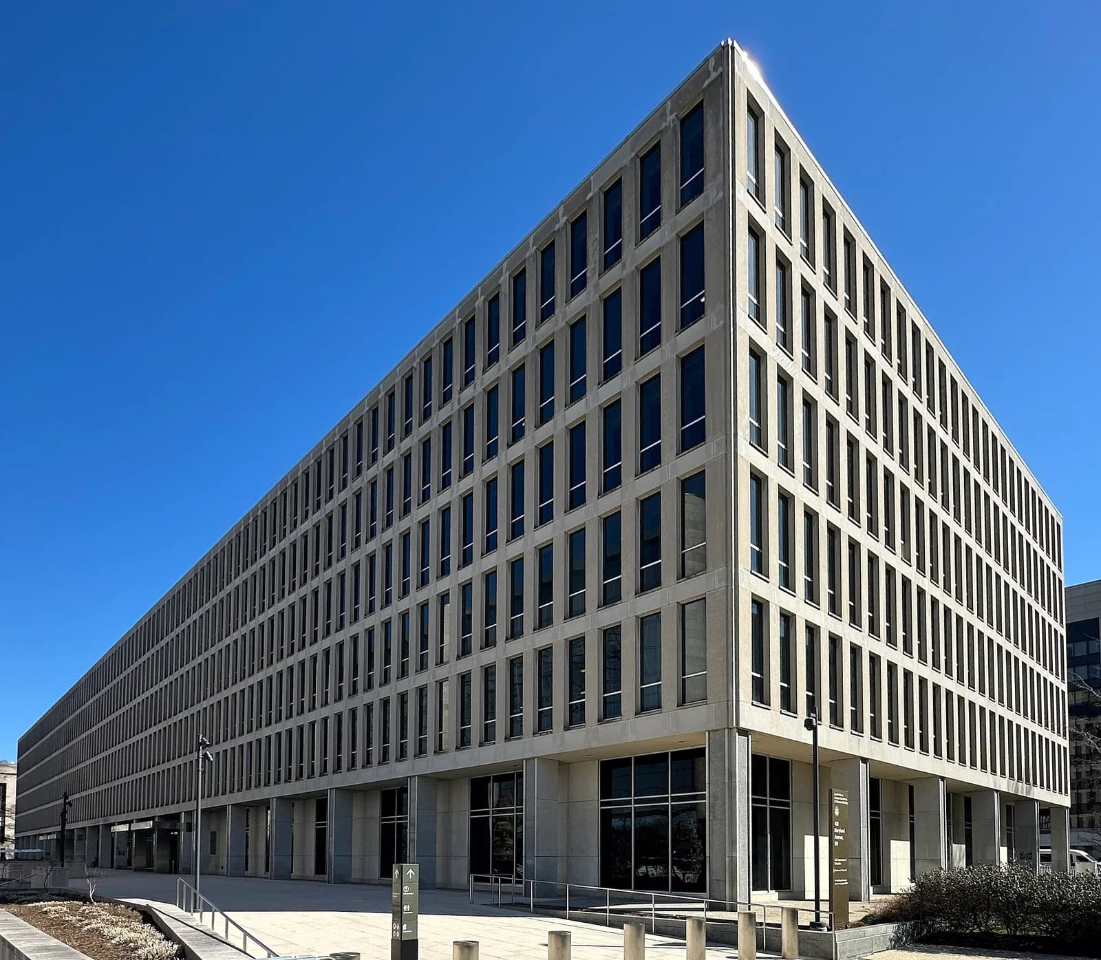 A large, modern, multi-story building with a grid of rectangular windows is shown from a low-angle perspective against a clear blue sky. The building's light-colored facade extends into the distance, creating a strong sense of depth.