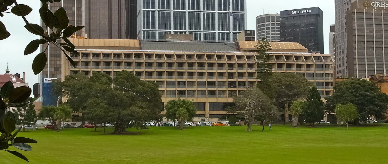 A large, multi-story building with a grid of balconies is visible behind a green lawn with trees. Other taller buildings rise in the background, and a few people are on the lawn.