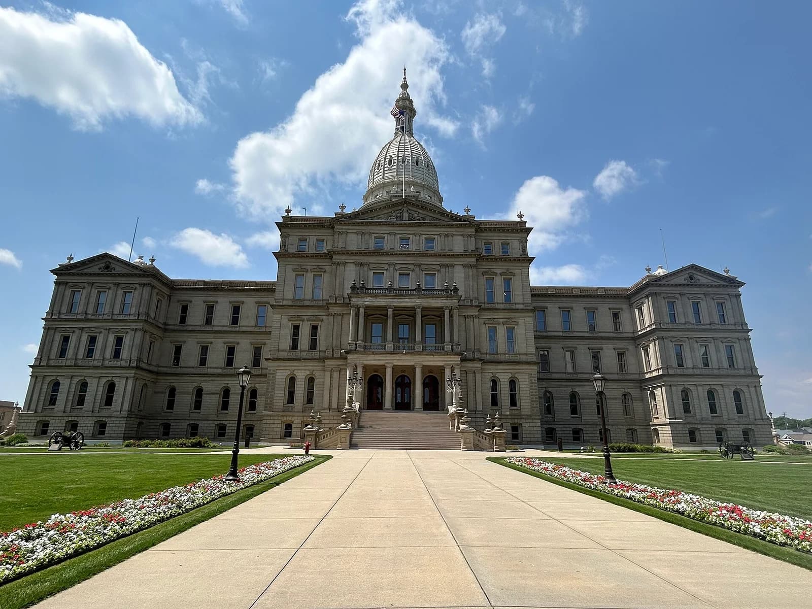 A grand, light-colored stone building with a large dome and columns stands under a blue sky with white clouds. A wide concrete path leads up to the building, flanked by green lawns and flower beds.