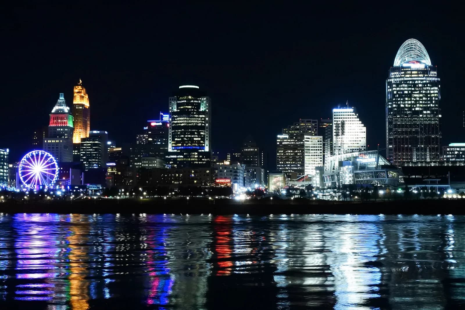 A nighttime cityscape shows numerous illuminated buildings, including a large Ferris wheel and a skyscraper with a domed top, reflected in the dark water of a river in the foreground.