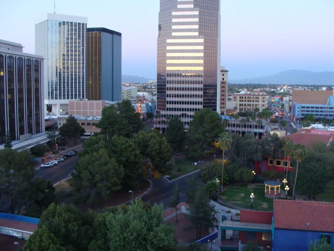 An elevated view of a city at dusk shows several tall buildings, a park with trees and a gazebo, and a street with cars. Distant mountains are visible under a pale sky.