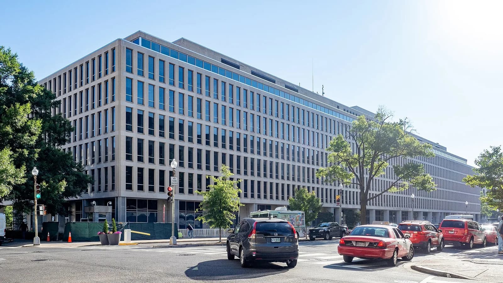 A large, modern building with many rectangular windows is seen from a street corner. Cars are visible on the road in front of the building, and trees line the street.