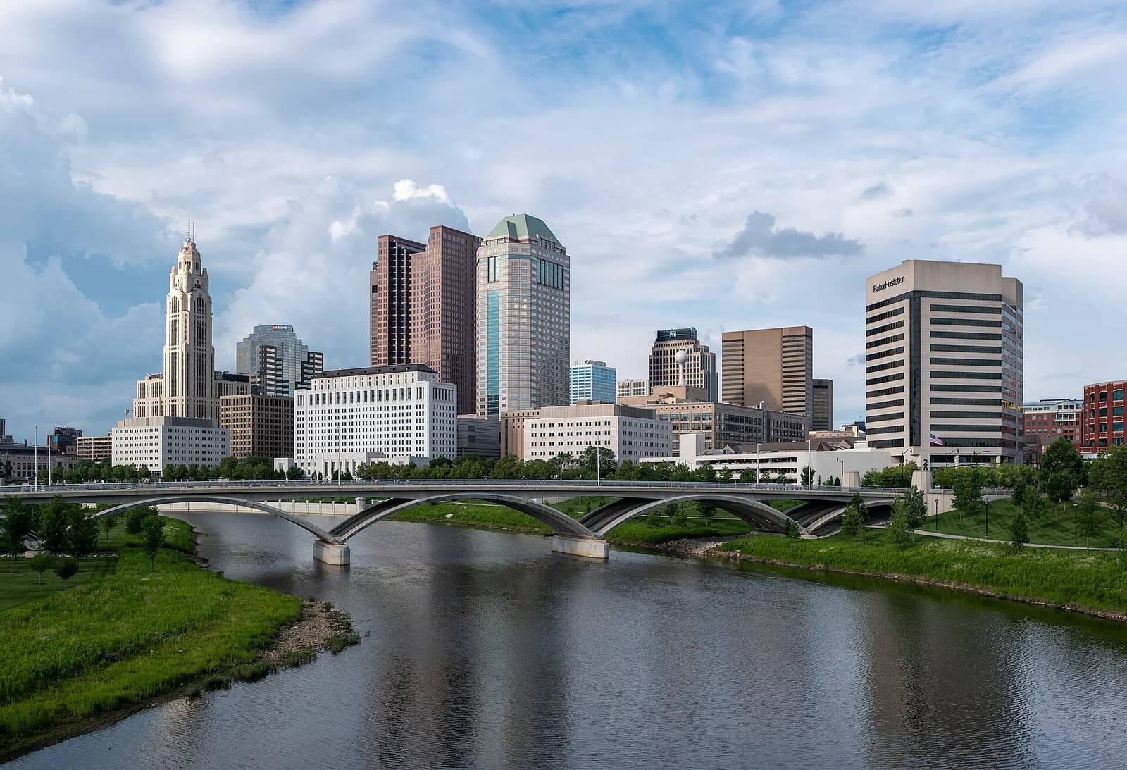 A cityscape features a river in the foreground with a bridge spanning it, leading to a cluster of diverse buildings under a partly cloudy sky. The buildings include a prominent tall, light-colored skyscraper with an ornate top, several modern glass and concrete structures, and a white building with many windows.